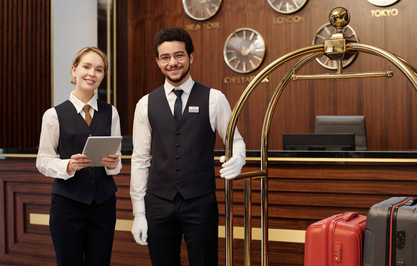 TIMELESS Hotelpersonal: Rezeptionistin mit Tablet und Page mit weißen Handschuhen an einem Gepäckwagen in einer eleganten Hotellobby mit Weltzeituhren