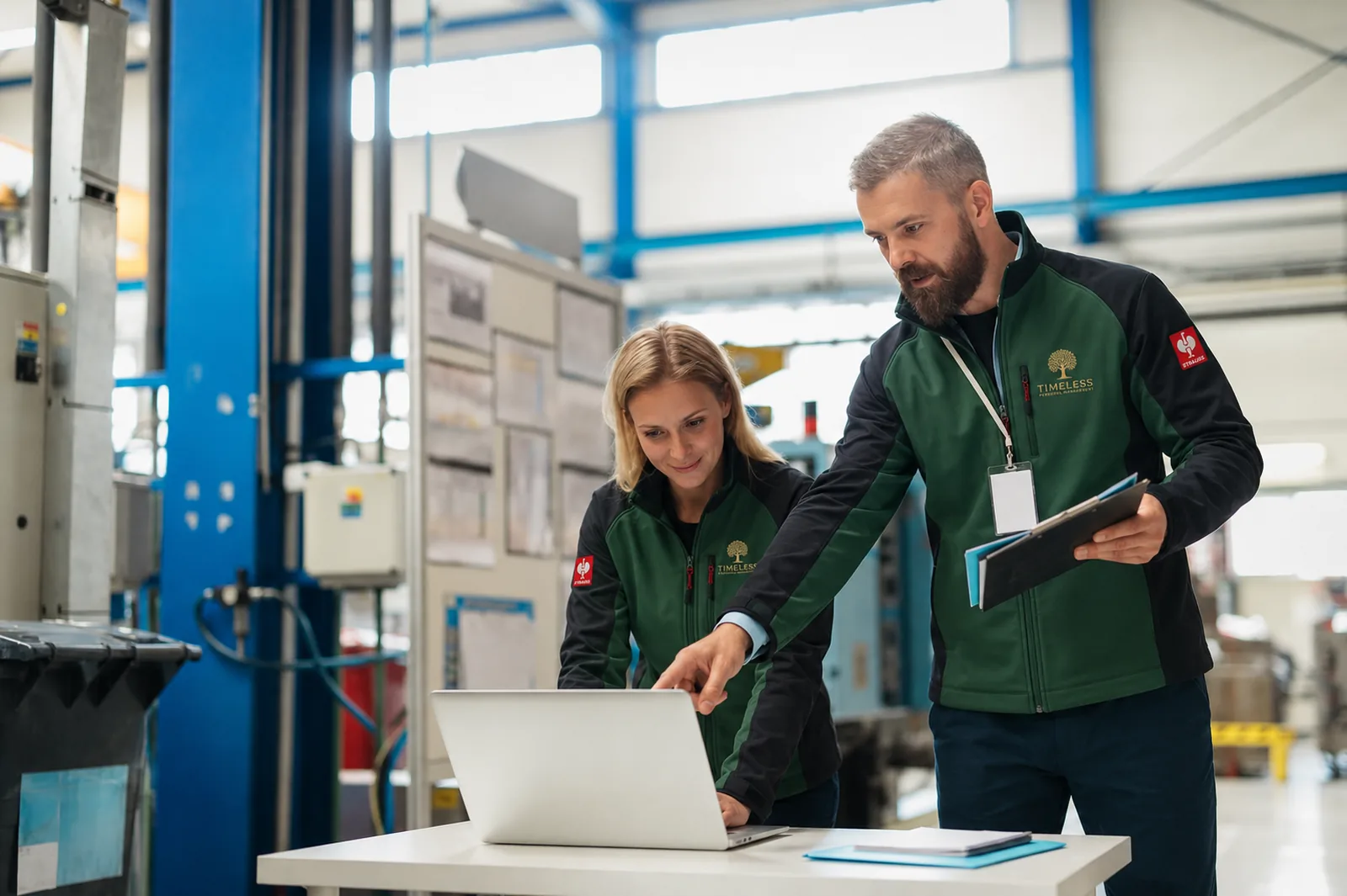 TIMELESS Team in grüner Softshell-Jacke bespricht Auftrag am Laptop in Werkstatt- und Logistikumgebung